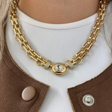 Gold chain necklace with a central gemstone worn by a person in a brown coat.