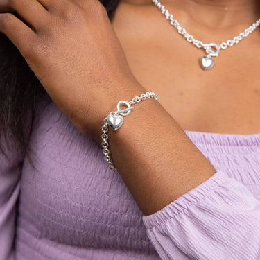 Silver bracelet with heart charm on a wrist, wearing a light purple top.