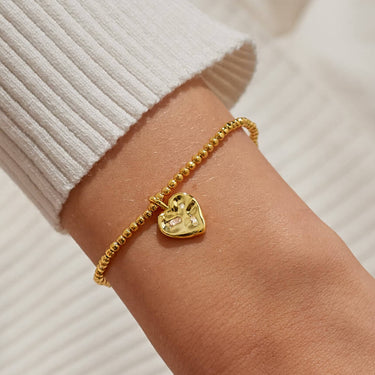 Gold bracelet with a heart charm on a wrist against a neutral background