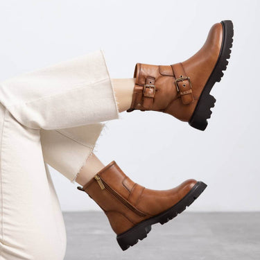 Brown leather boots with buckles worn by a person sitting on a light gray floor.
