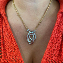 A close-up of a person's chest wearing a gold chain necklace with a polished silver pendant made of interlocking curved loops, styled with a bright orange knit top.
