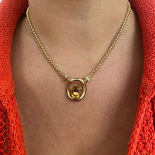 A gold chain necklace with a central gold ring and a round yellow gemstone pendant, worn on a model's neck over a bright orange knit top.