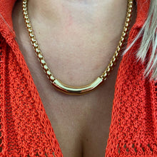 A close-up of a gold-toned curved bar necklace worn by a model wearing a bright coral-red knit top.