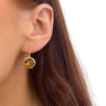A close-up of a woman's ear wearing a gold hoop earring with a round amber-colored stone pendant, against a clean white background.