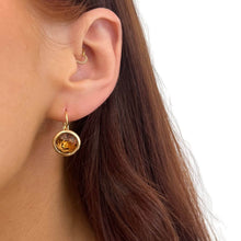 A close-up of a woman's ear wearing a gold hoop earring with a round amber-colored stone pendant, against a clean white background.