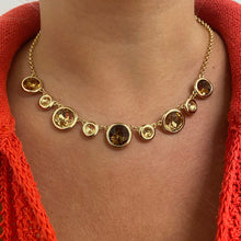 A gold chain necklace with multiple round amber-colored stones in bezel settings, worn by a person in a bright orange knit top.