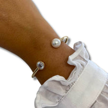 A close-up of a person's wrist wearing two delicate gold bracelets: one with a single white pearl and the other with a clear round gemstone, paired with a white ruffled sleeve against a white background.