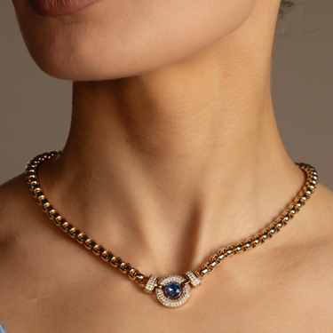 Gold necklace with a blue gemstone on a person's neck against a neutral background