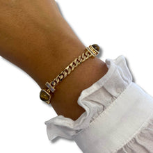 A gold-toned chain bracelet with rectangular links and oval brown gemstone accents, shown worn on a person's wrist above a white ruffled sleeve.