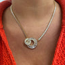 A close-up of a person's neck and chest wearing a gold chain necklace with a double interlocking spiral pendant set with clear baguette and round stones, styled with a bright orange knit top.