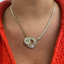 A close-up of a person's neck and chest wearing a gold chain necklace with a double interlocking spiral pendant set with clear baguette and round stones, styled with a bright orange knit top.