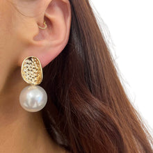 A close-up of a model's ear wearing a gold drop earring with a textured oval top and a dangling white pearl, against a clean white background.