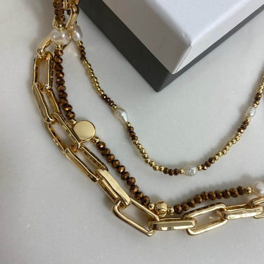 A layered necklace set featuring a chunky gold paperclip chain, a delicate gold chain with brown beads and white pearl accents, and a thin gold chain with small brown beads, displayed on a light gray surface next to a white and black box.