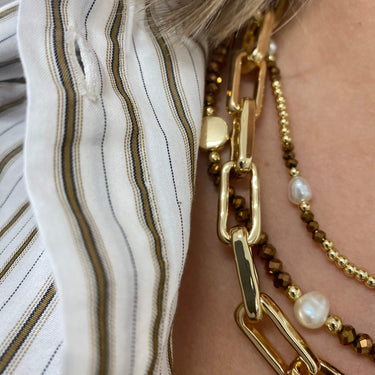Close-up of layered necklaces worn on a person's neck, featuring a chunky gold chain, a beaded brown and gold chain, and a pearl necklace, next to a white and brown striped shirt.
