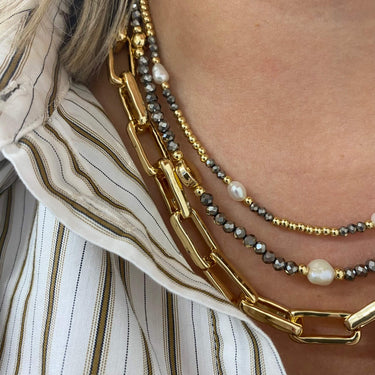 Close-up of a person's neck wearing three layered gold necklaces: a chunky link chain, a beaded chain with gray faceted beads and white pearls, and a delicate chain with a larger white pearl.