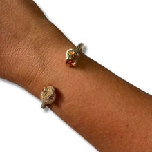 Two gold knot-shaped rings, one with pavé diamonds and one with a red gemstone, shown on a person's wrist against a white background.