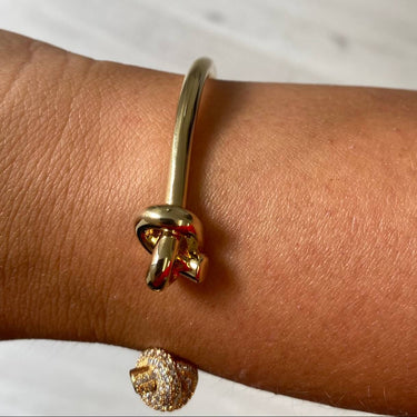A gold-toned metal cuff bracelet with a knotted detail, shown worn on a person's wrist. A second small gold-toned crystal-encrusted charm is dangling below the wrist.