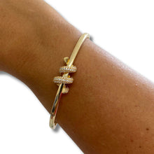 A gold bangle bracelet with a twisted knot design paved with small clear stones, shown worn on a person's wrist against a white background.