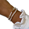 A close-up of a person's wrist wearing two pearl beaded bracelets with gold starburst charms, styled with a white ruffled sleeve on a plain white background.