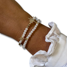 A close-up of a person's wrist wearing two pearl beaded bracelets with gold starburst charms, styled with a white ruffled sleeve on a plain white background.