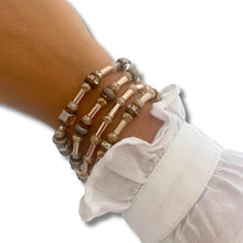 A close-up of a person's wrist wearing three stacked beaded stretch bracelets in neutral earthy tones, paired with a white ruffled shirt cuff against a plain white background.