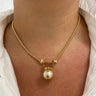 A close-up of a person's neck and chest wearing a gold chain necklace with a rectangular gold frame holding a single pearl pendant, styled with a white collared shirt.