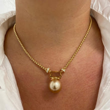 A close-up of a person's neck and chest wearing a gold chain necklace with a rectangular gold frame holding a single pearl pendant, styled with a white collared shirt.
