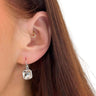 A close-up of a person's ear wearing a small silver hoop earring in the cartilage and a dangling silver hook earring with a faceted clear gemstone, against a white background.