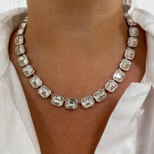 A close-up of a model wearing a silver-toned collar necklace with rectangular clear faceted gemstones, styled with a white button-down shirt.