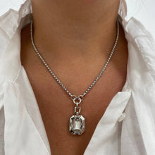 A close-up of a person wearing a silver chain necklace with a rectangular clear faceted pendant, styled with an open white blouse.