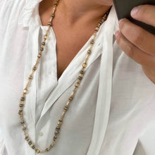 A close-up of a person wearing a long beaded necklace over a white button-up shirt, holding a phone in one hand.