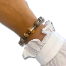 A close-up of a person's wrist wearing a beaded bracelet with gray and gold beads, a faceted clear crystal cube, and a textured gold bead, styled with a white ruffled sleeve.