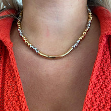 A close-up of a person's neck and upper chest wearing a two-tone gold and silver curved necklace over a bright coral-red knit top.