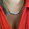A close-up of a person wearing a gold chain necklace with a polished silver curved bar at the center, styled with a bright coral orange knit top.