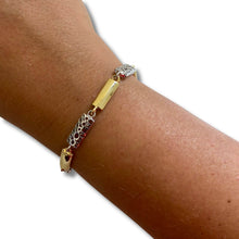 A gold and silver bracelet worn on a person's wrist, featuring a central polished gold bar, textured silver links, and a small red gemstone accent.