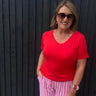 Woman wearing a red shirt and pink striped pants standing against a dark wooden wall.