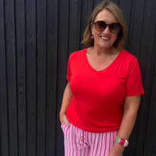 Woman wearing a red shirt and pink striped pants standing against a dark wooden wall.