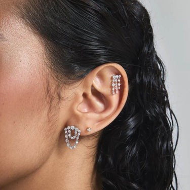 A close-up of a woman's ear wearing a silver-toned ear cuff with clear stones and a matching small stud earring, against a light gray background.