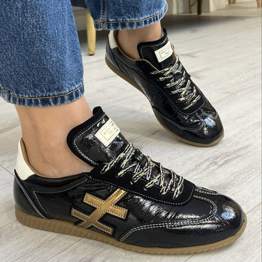 A person wearing black glossy sneakers with gold accents and white laces, paired with blue jeans, standing on a light wood floor in an indoor setting.