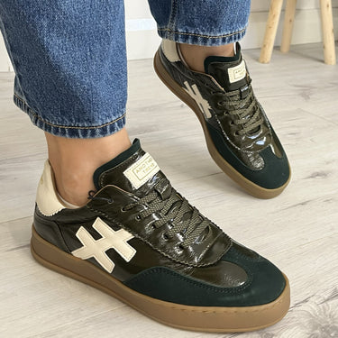 A person wearing dark green and black low-top sneakers with cream-colored cross-shaped side accents, paired with blue jeans in a home setting with a light wood floor and a wicker basket planter in the background.