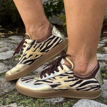 A pair of metallic gold and black zebra-print sneakers with brown laces, shown being worn outdoors on a stone surface.