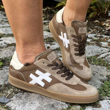 A person wearing brown suede and canvas low-top sneakers with white graphic cross logos, standing on a stone path outdoors.