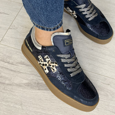 A person wearing dark navy low-top sneakers with leopard-print side accents, gray laces, and gum soles, paired with blue jeans on a light wood floor.