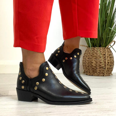 Black ankle boots with gold studs worn with red pants on a light wooden floor.