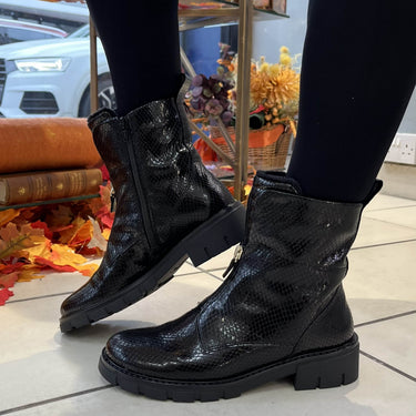 Black snakeskin-textured ankle boots with chunky lug soles, shown on a model wearing black leggings in a store setting.