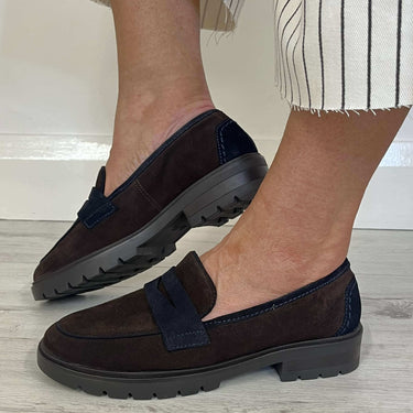 A person wearing dark brown suede slip-on loafers with navy blue trim and a chunky black sole, paired with white pants featuring thin black vertical stripes, standing on a light gray wood floor.