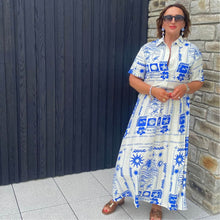 Woman wearing a blue and white patterned dress standing against a black wall and stone wall.