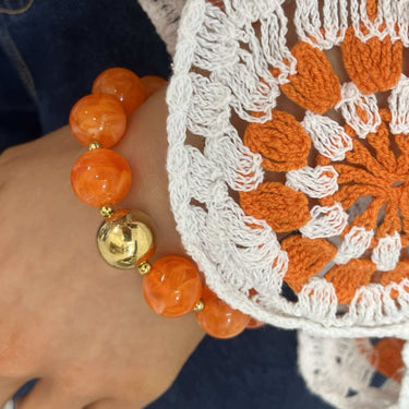Close-up of orange bracelet with gold charm on wrist wearing a white lace garment.