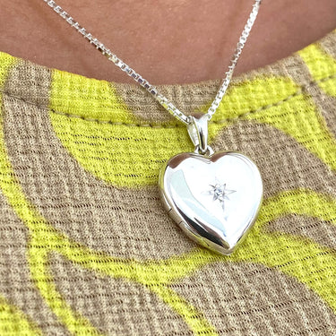 Silver heart-shaped locket with a diamond on a chain against a textured fabric background.