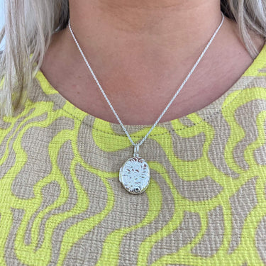 Silver necklace with a oval locket pendant worn by a person against a patterned fabric background